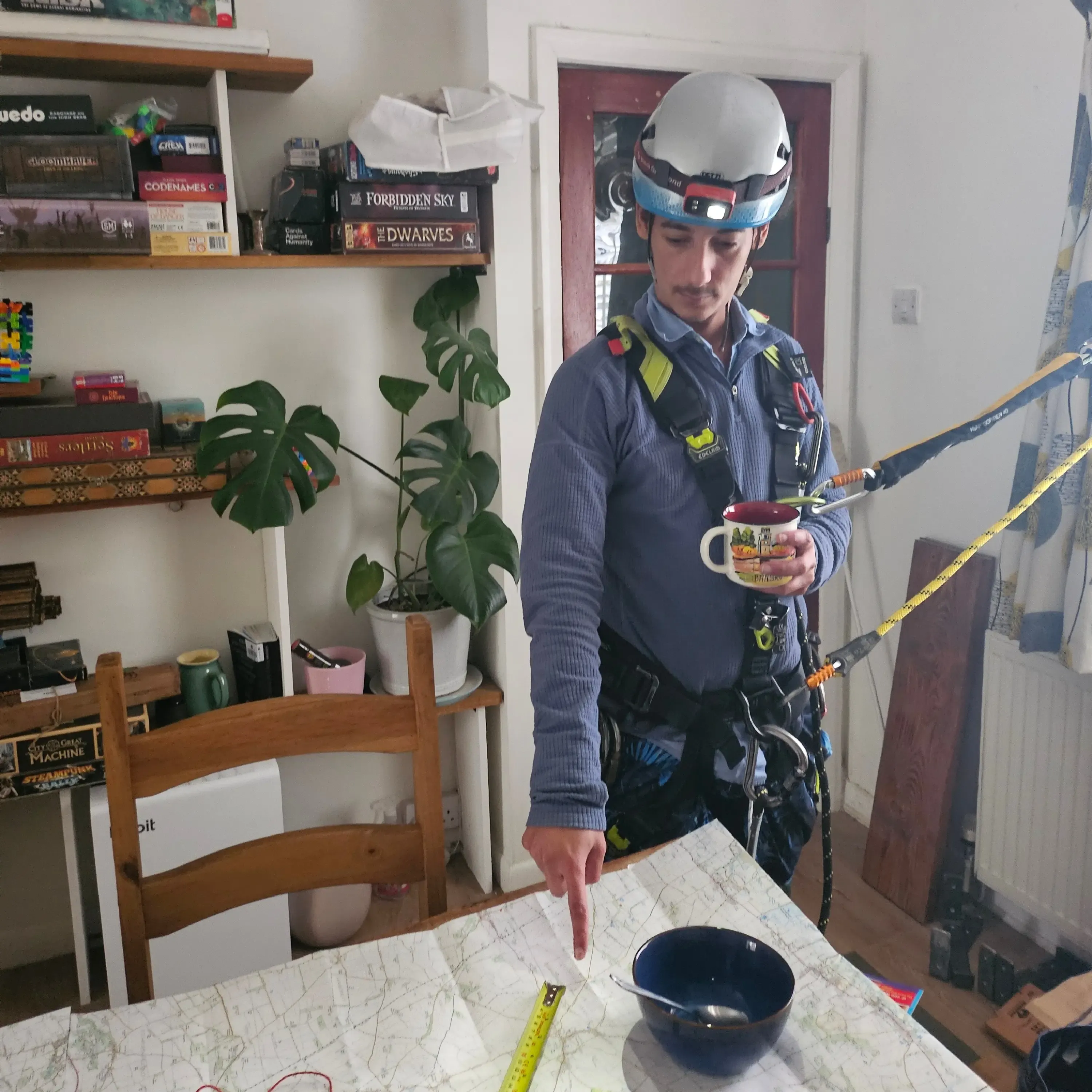 QRidian co-founder Bernie wearing full climbing gear indoors, pointing clinically at a map while holding a tea mug. Captures the team's blend of technical ability and humour.
