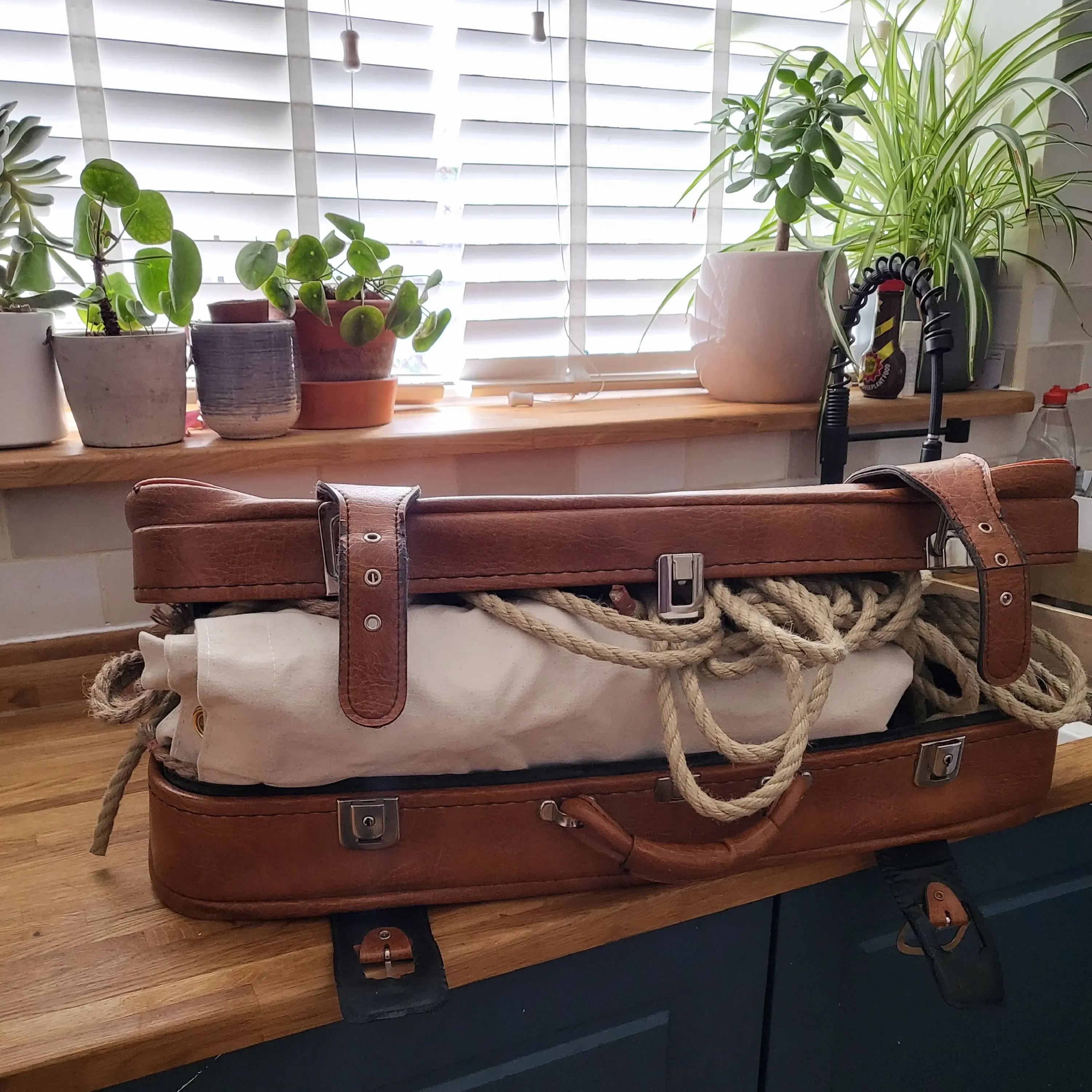 An open vintage brown leather suitcase on a wooden kitchen counter, packed tightly with rolled cream canvas and thick rope. The image highlights the handmade, portable elements of the physical gameplay kits.
