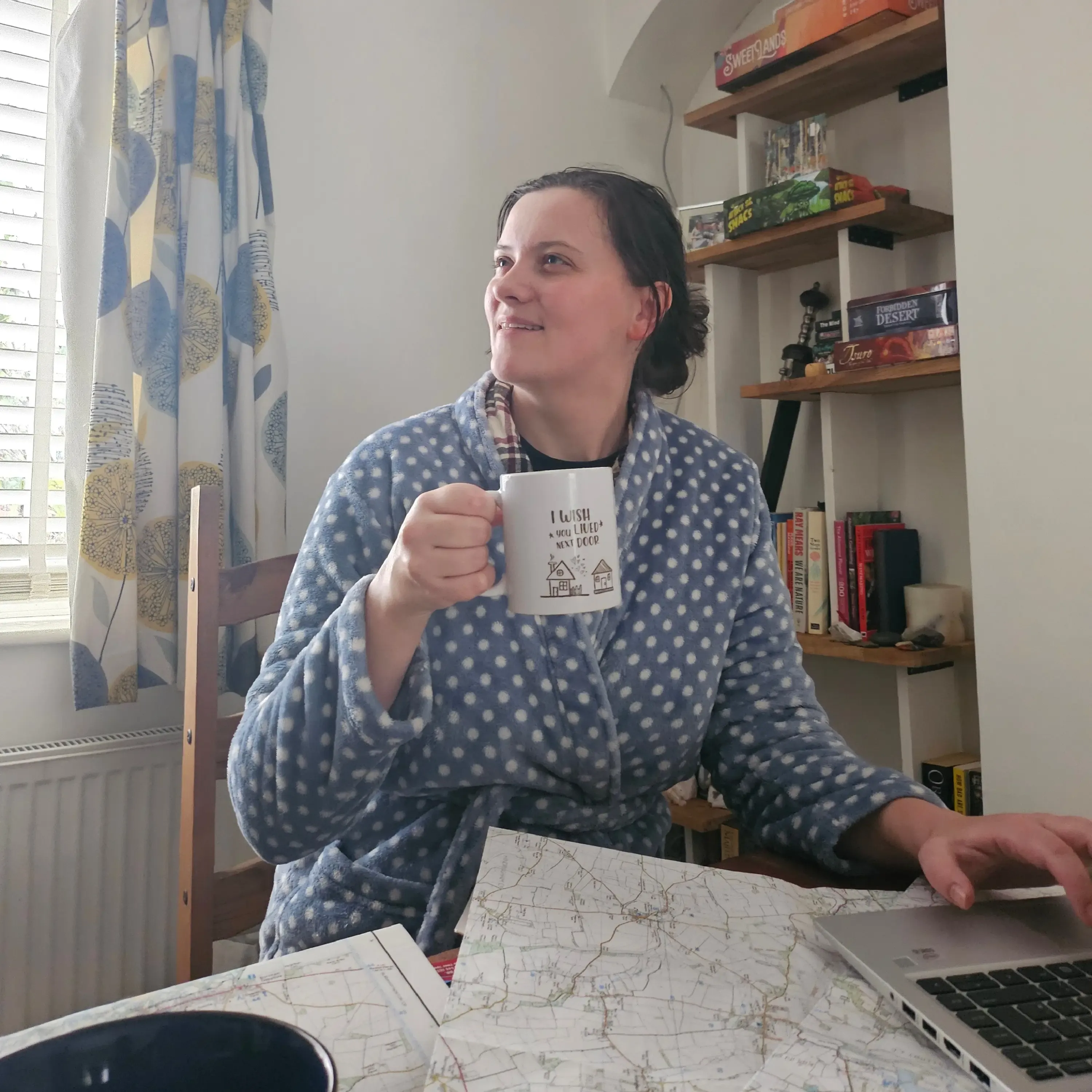 QRidian co-founder Molly smiles warmly at a table covered in maps and a laptop, wearing a cozy polka-dot dressing gown and holding a mug