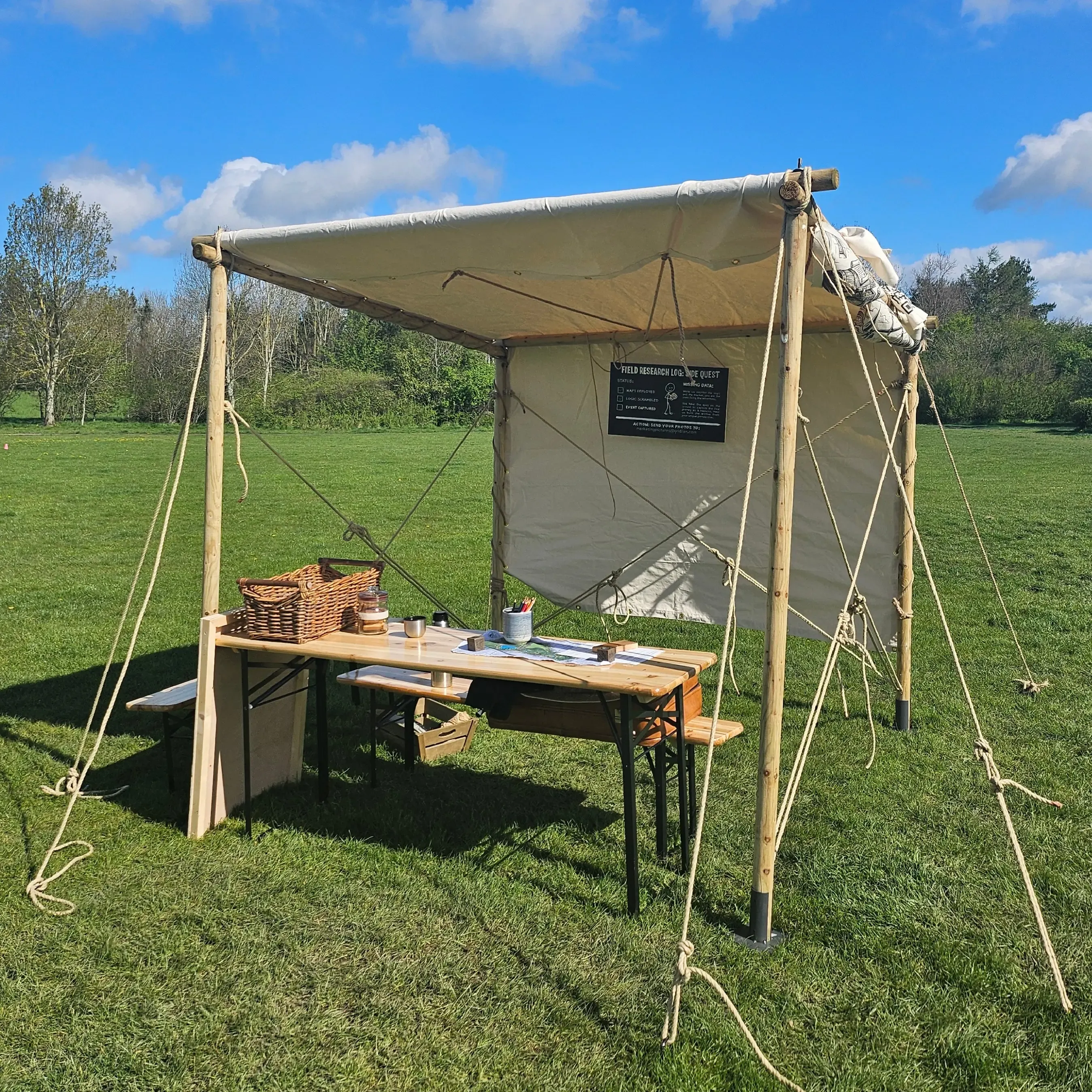 A portable canvas pop-up tent on a grass field, featuring a wooden table with maps, pencils, and a wicker basket. A sign reads 